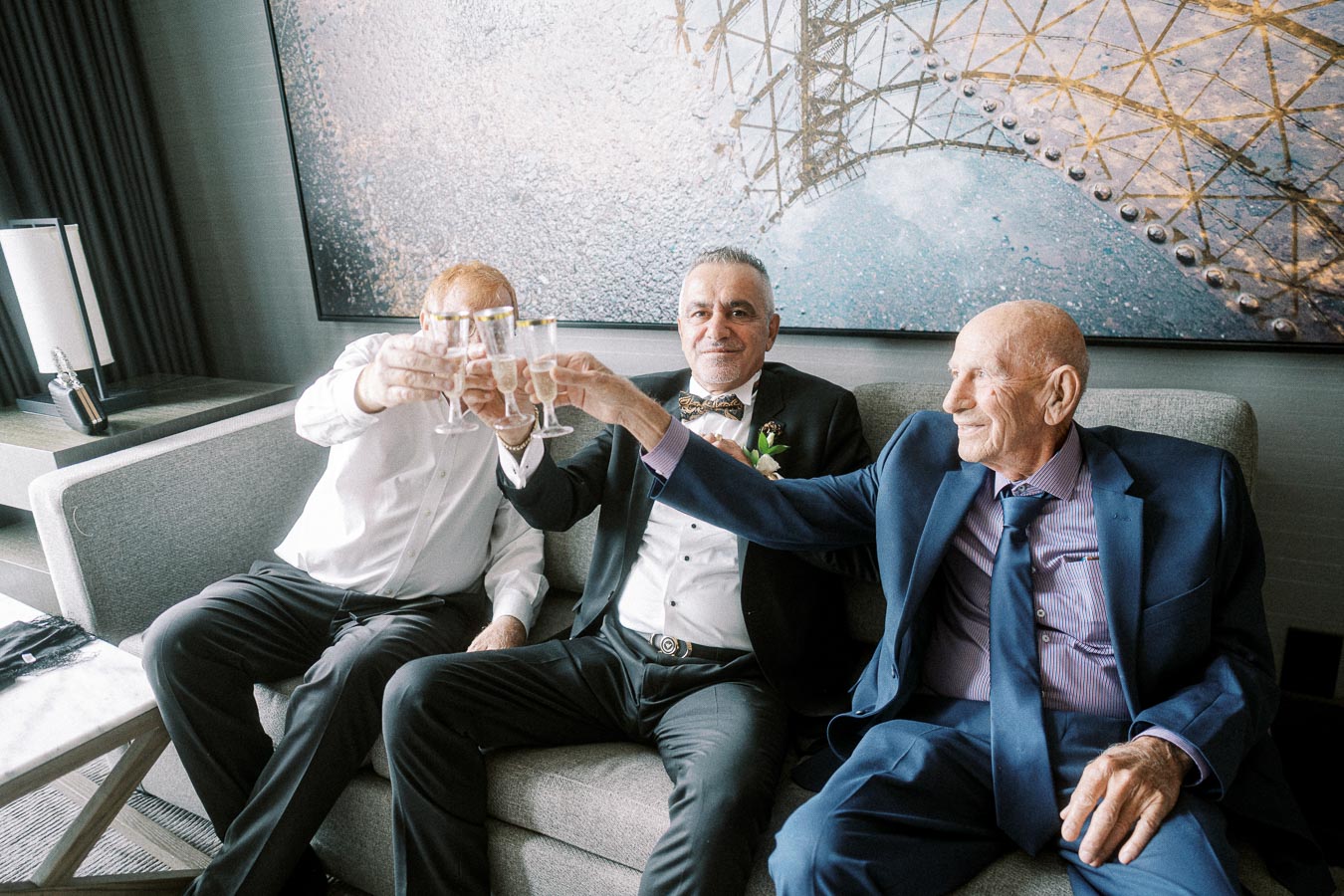 Three elderly men in formal attire making a toast with champagne glasses, seated on a sofa in a sophisticated living room