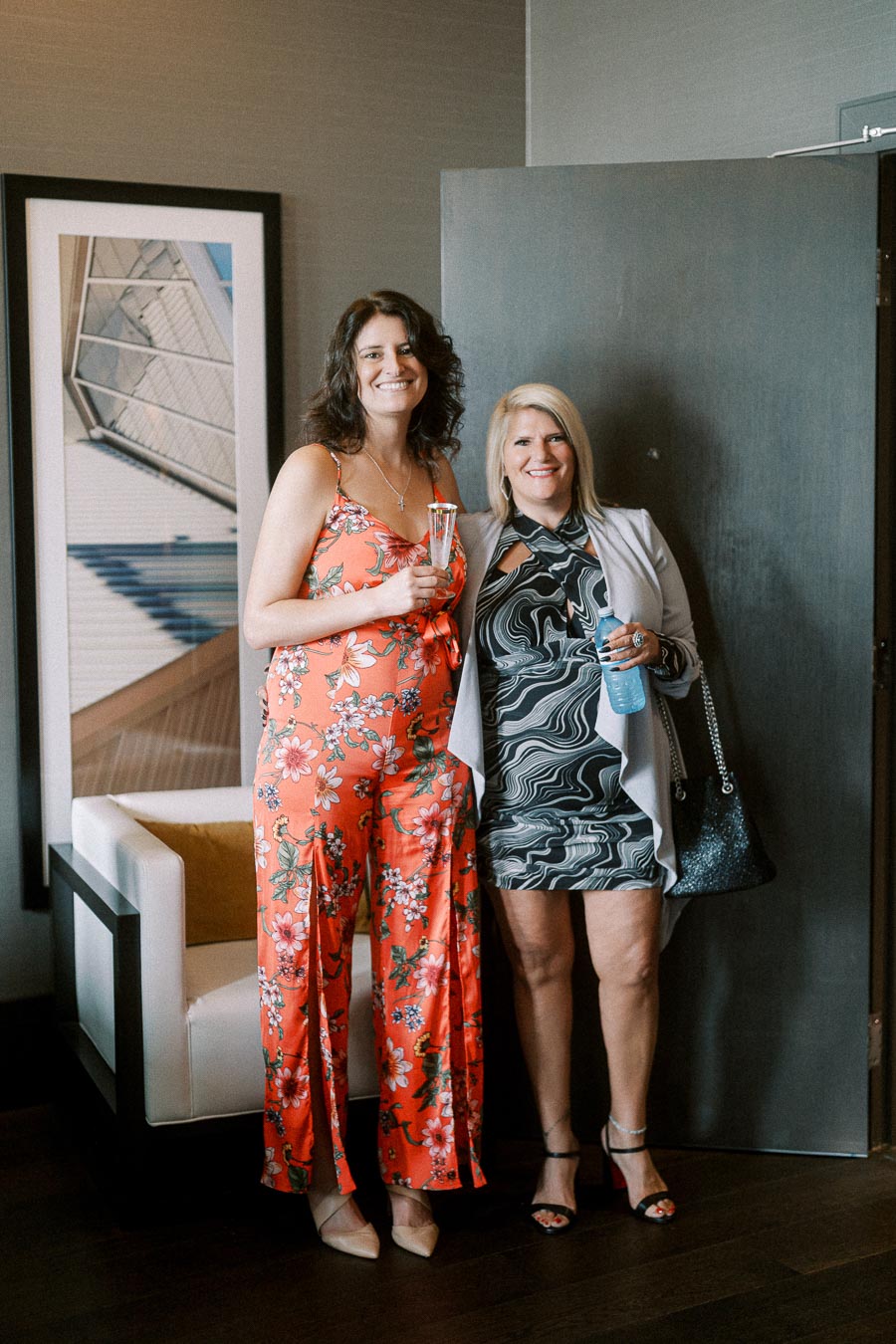 Two women in stylish outfits enjoying a social gathering, one in a floral jumpsuit holding a drink and the other in a