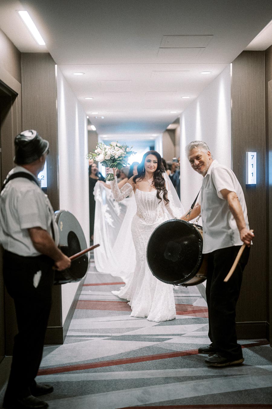 Bride in a white gown holding a bouquet, walking down a hallway with drummers creating a festive atmosphere.