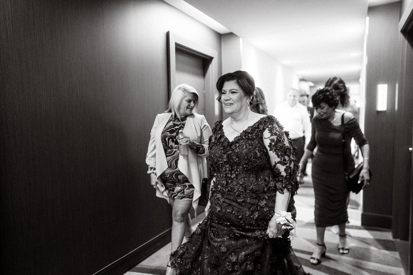 A woman wearing an elegant lace gown walks down a stylish hallway, accompanied by a group of well-dressed guests.