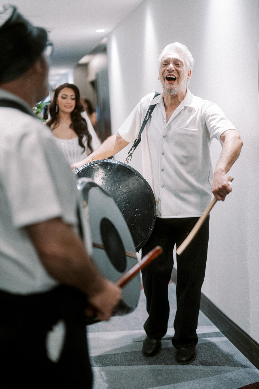 Elderly man joyfully playing a drum in a hallway, wearing a white shirt and black pants. Blurred figures and another drummer