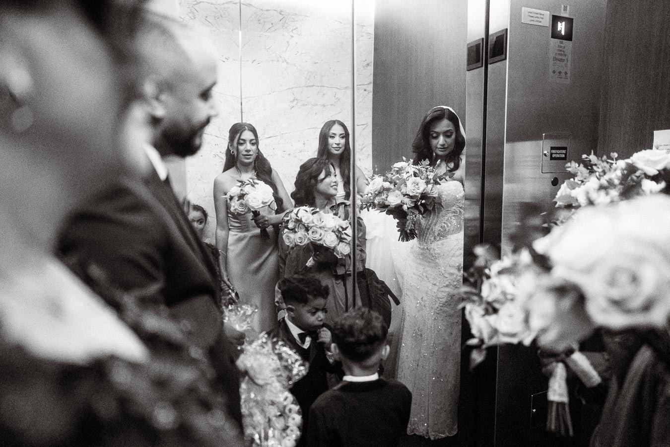 Black and white image of a bride holding a bouquet, surrounded by bridesmaids and children in an elevator, capturing a