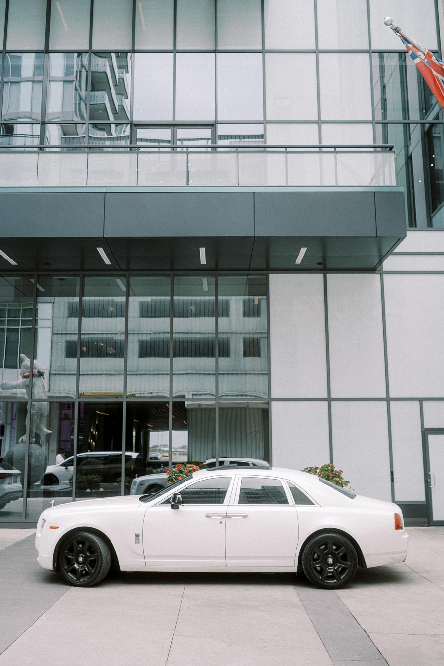 Luxury white Rolls-Royce parked in front of a modern glass building with sleek black rims and reflective windows,