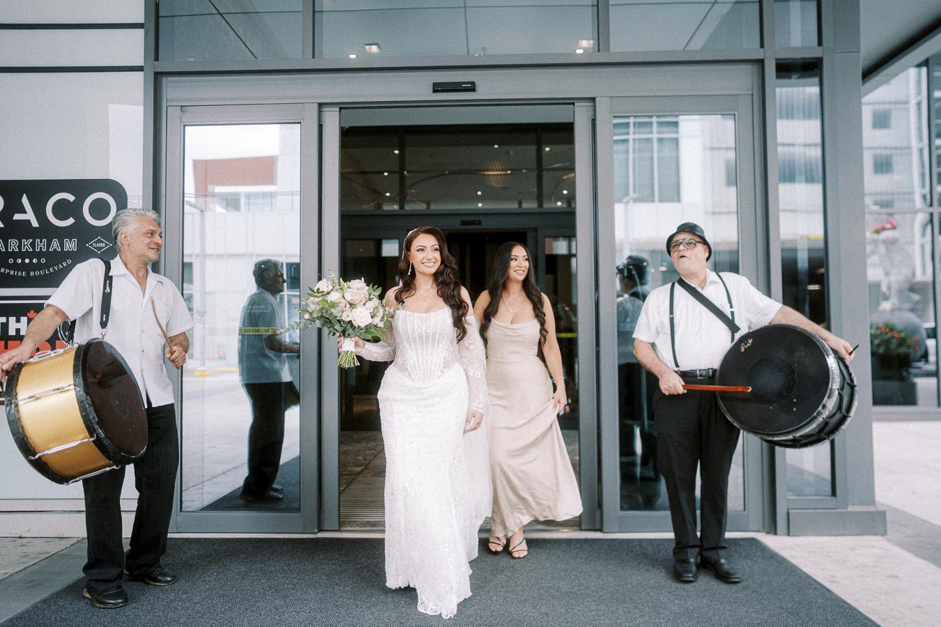 A bride and bridesmaid exiting a building with flower bouquet, accompanied by two drummers in celebration, with a modern