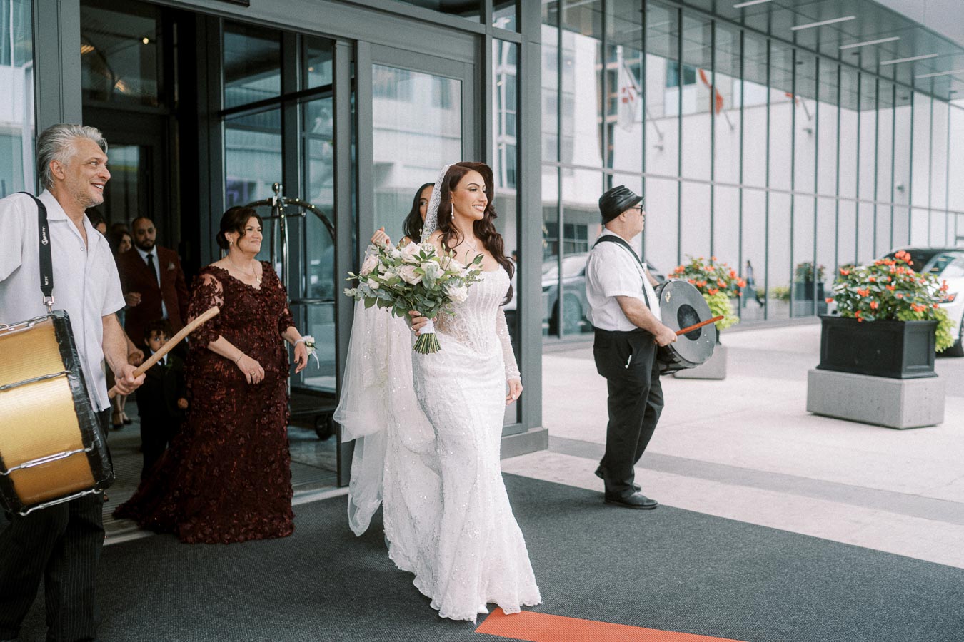 A bride in a lace wedding dress holding a bouquet of flowers exits a building, accompanied by musicians and guests in formal