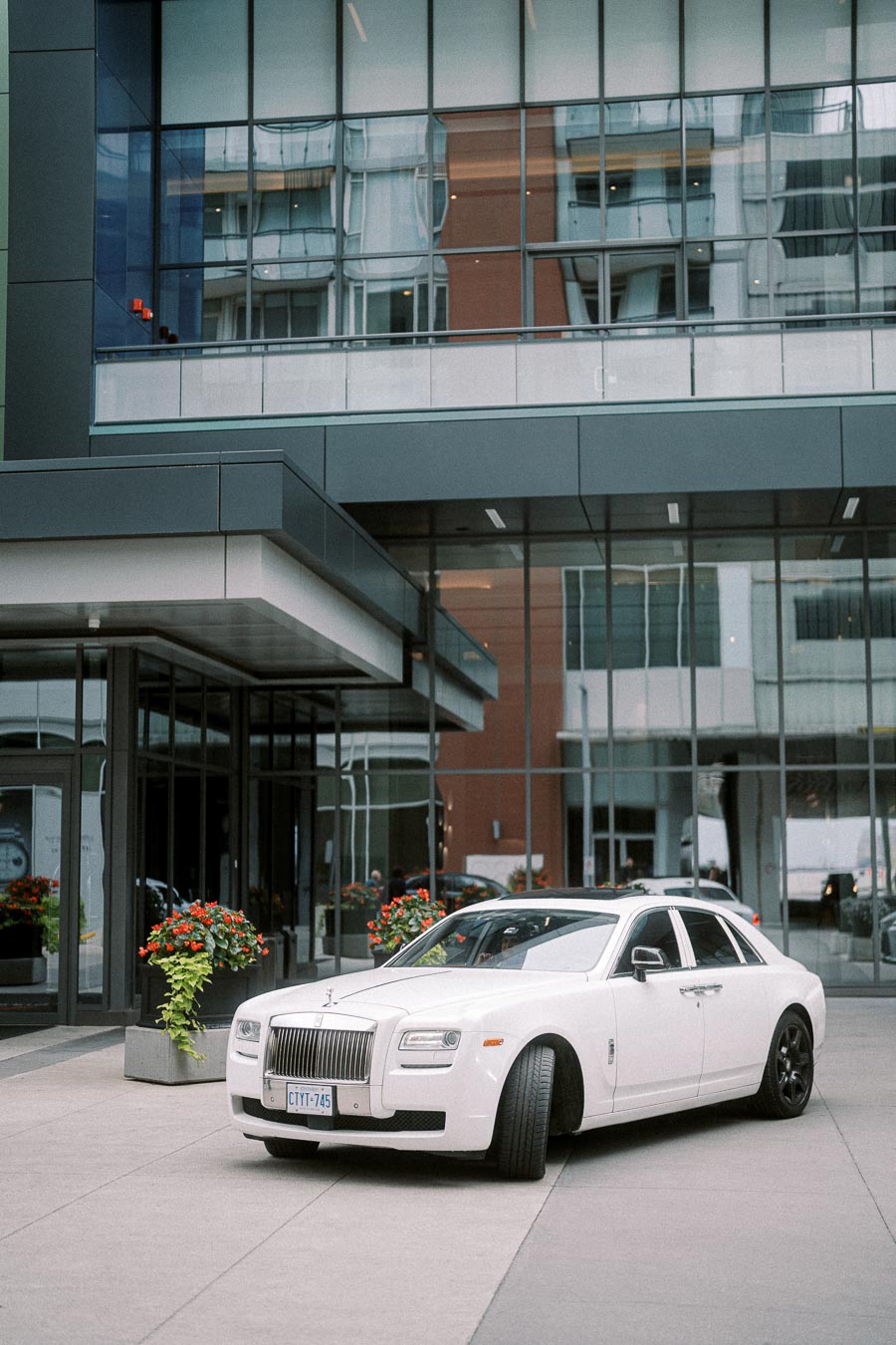 Luxury white Rolls-Royce parked in front of a modern glass building with red flowers, showcasing elegance and style in an