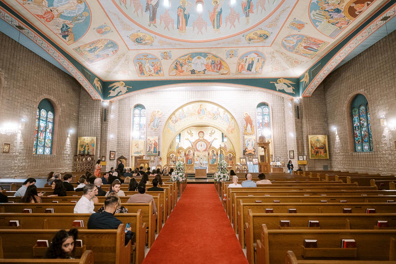 Interior of an ornate church with a red carpet aisle, stained glass windows, colorful ceiling frescoes, and wooden pews