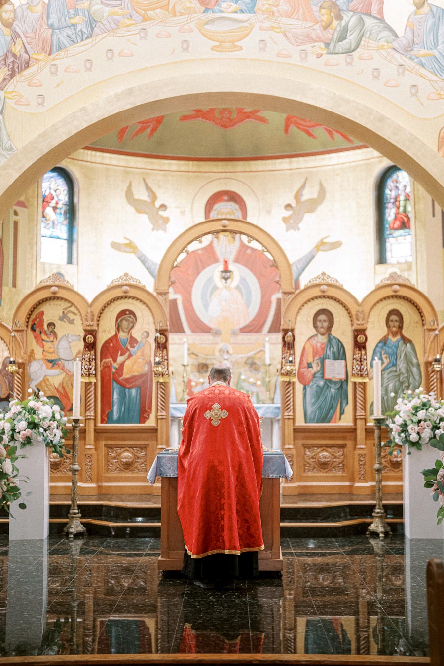 A priest in red vestments stands at an ornate altar in a richly decorated church interior featuring religious iconography