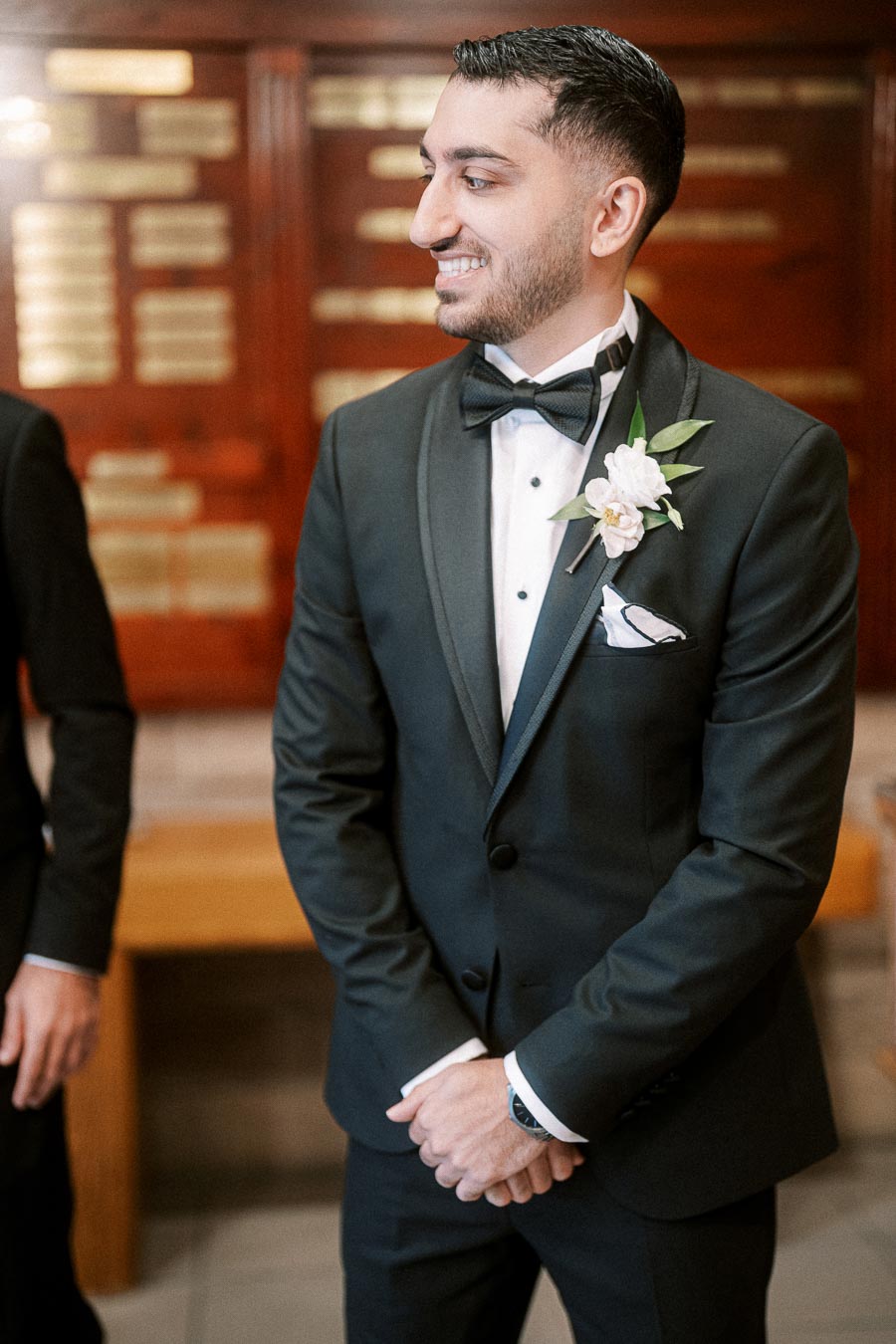 A groom in a black tuxedo with a white boutonniere, smiling and standing indoors against a blurred background of wooden