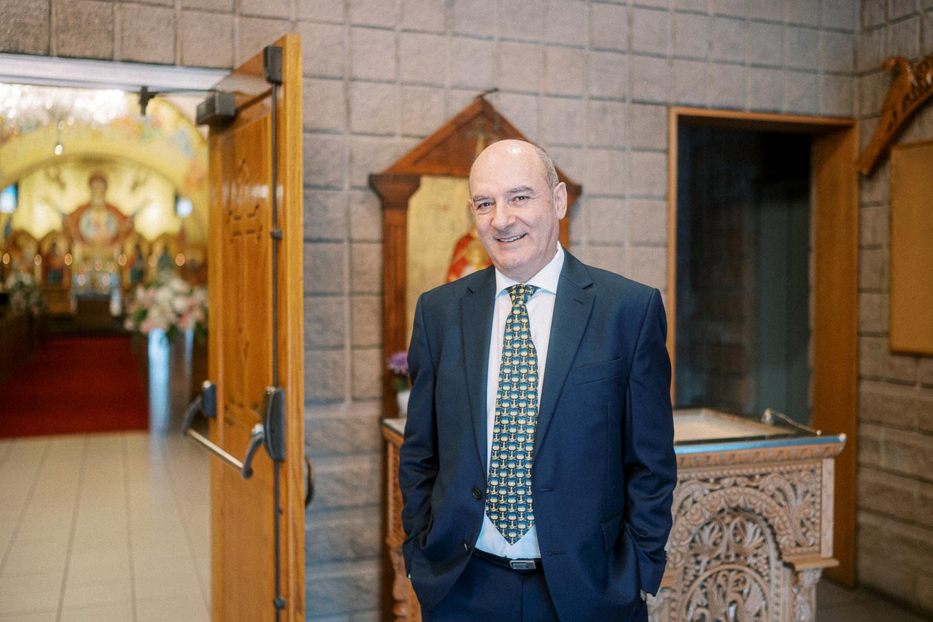 A man in a suit standing in the entrance of an ornate church, smiling at the camera, with decorative woodwork and religious