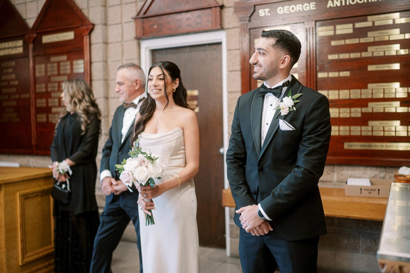 A bride and groom in formal attire, smiling and standing together indoors, accompanied by two elegantly dressed individuals,