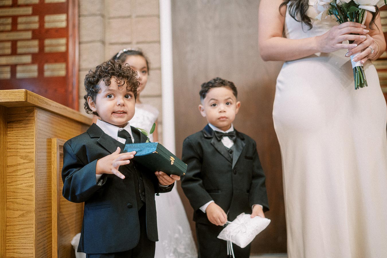 Cute young boys dressed as ring bearers holding a cushion and box at a wedding ceremony, standing near a woman in a white