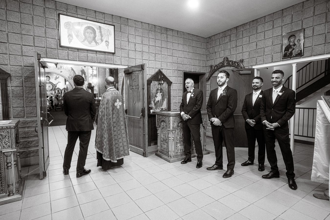A group of groomsmen in suits standing inside a church entrance, alongside a priest with a detailed robe, preparing for a