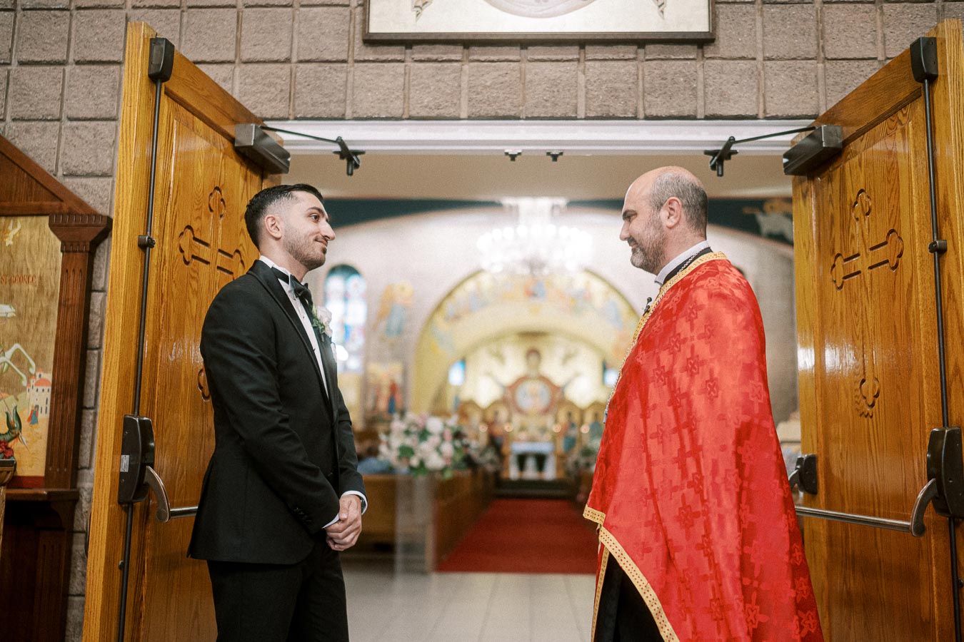 A groom in a black suit standing and smiling at a priest in red vestments inside a decorated church with open wooden doors