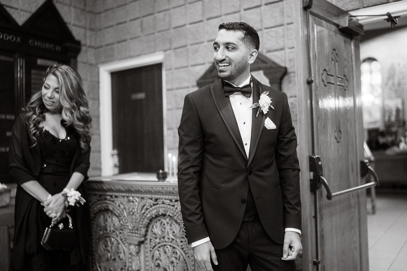 A smiling groom in a tuxedo stands next to a woman in a formal dress inside a church, capturing a joyful wedding moment.