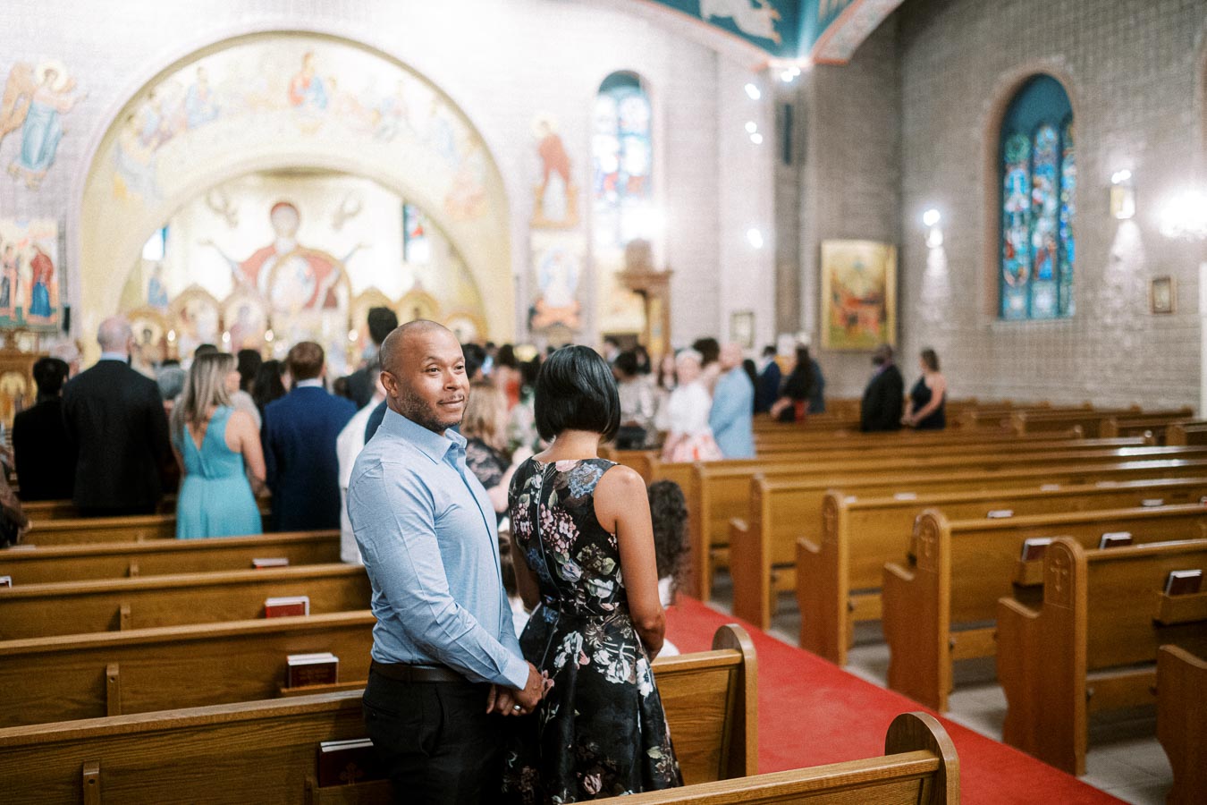 A couple standing in a beautifully adorned church with wooden pews and a large mural, surrounded by people during a ceremony