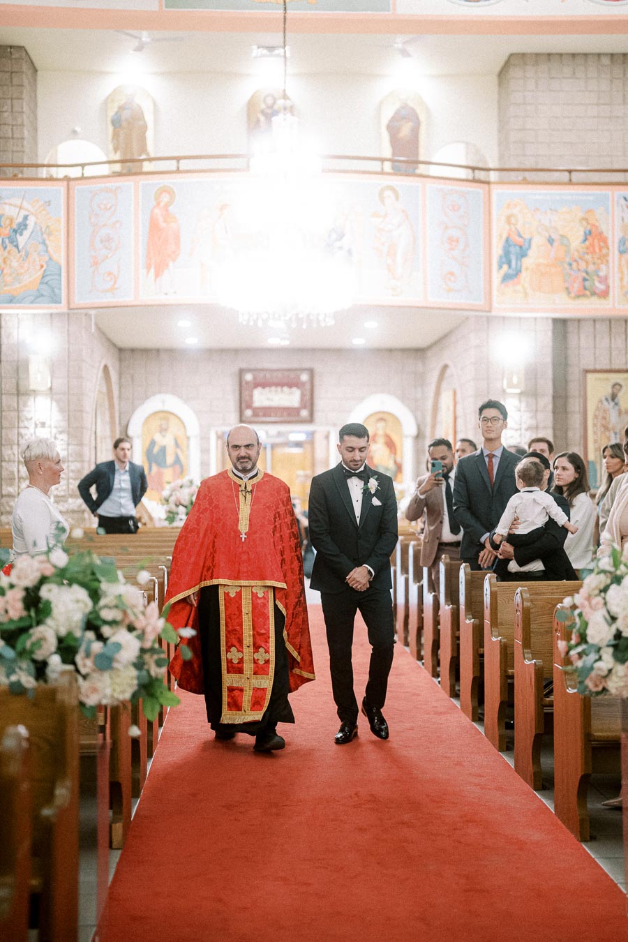Priest in traditional red vestment and groom walking down the aisle of a decorated church during a wedding ceremony,