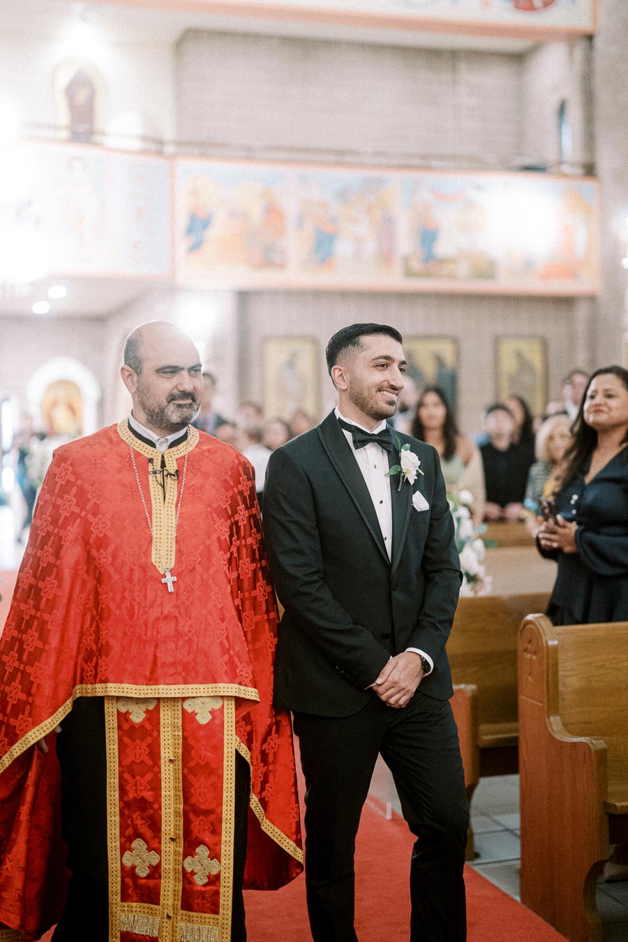 Wedding ceremony in a church with a priest in red robes standing beside a groom in a black tuxedo, surrounded by guests