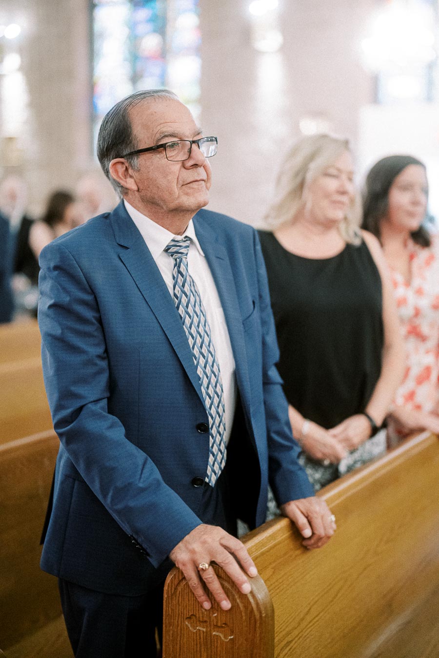Elderly man in a blue suit standing in a church pew during a wedding ceremony, with stained glass windows in the background.