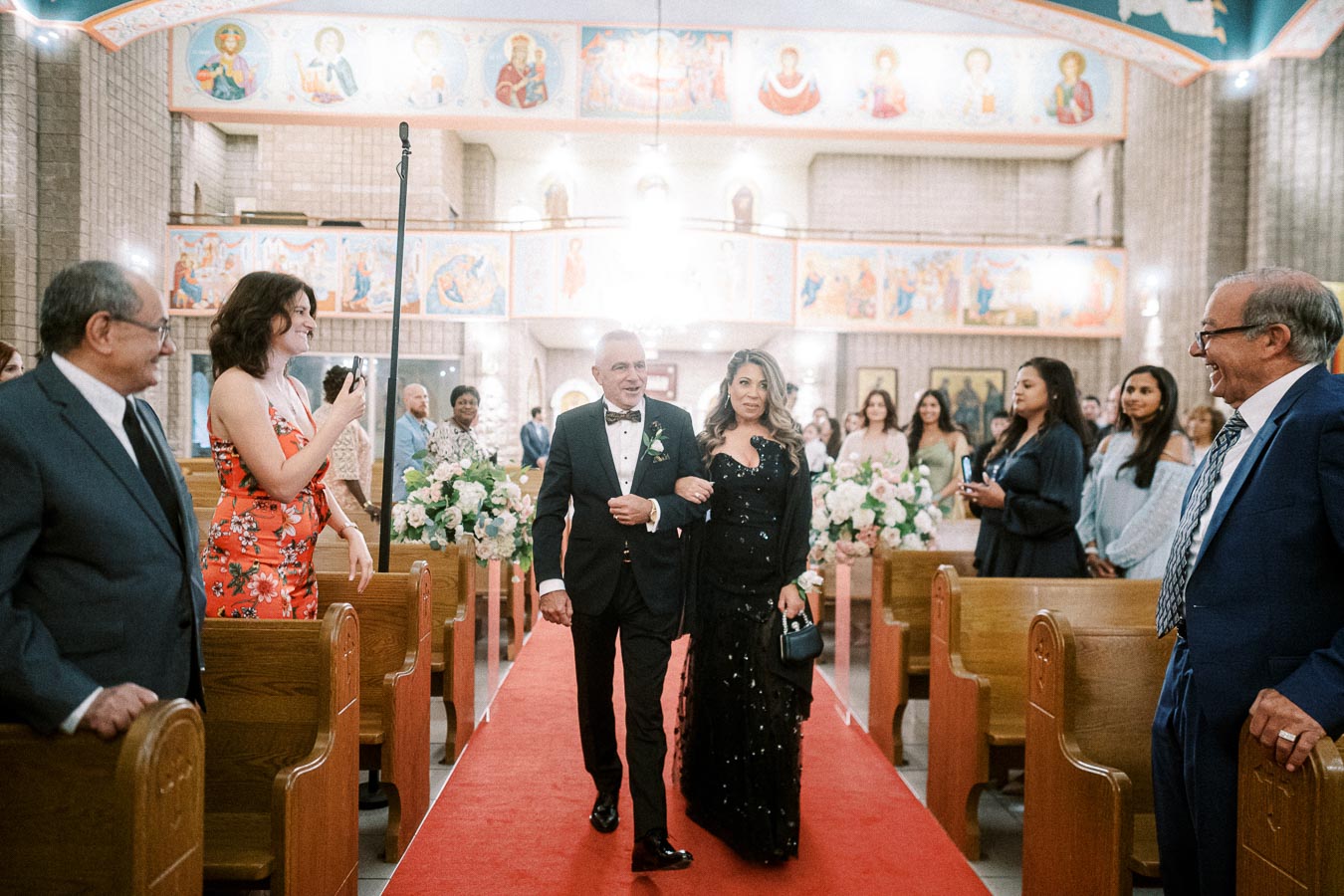 A couple elegantly dressed, walking down the aisle in a beautifully decorated church with guests looking on during a formal