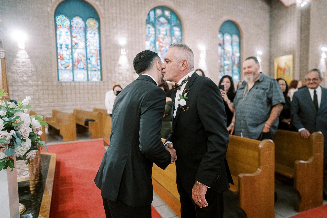 A heartfelt moment during a wedding ceremony in a church, featuring two men in suits shaking hands and sharing a warm