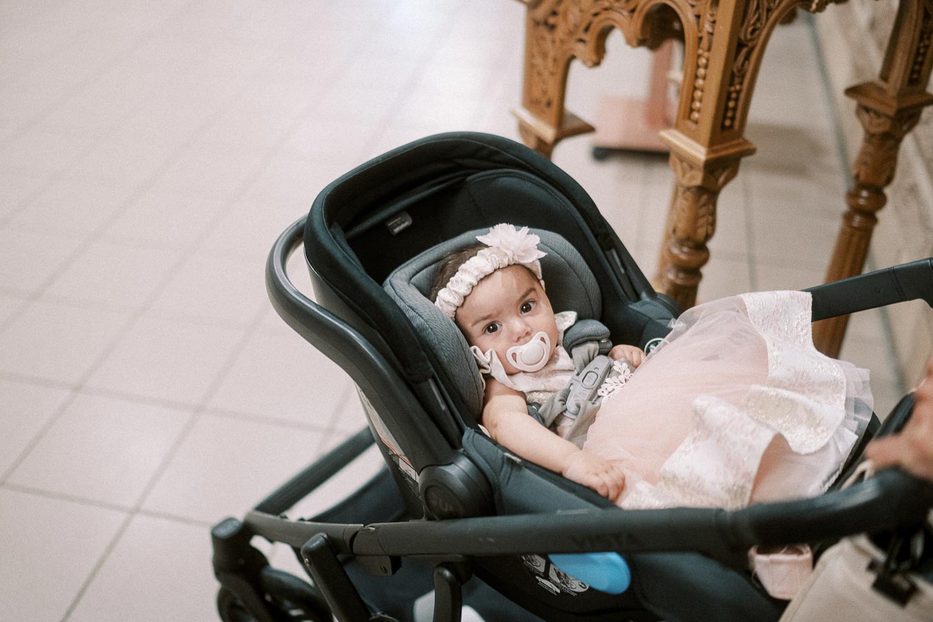 Cute baby in a stroller with a pacifier and a pink dress, showing a curious expression indoors.