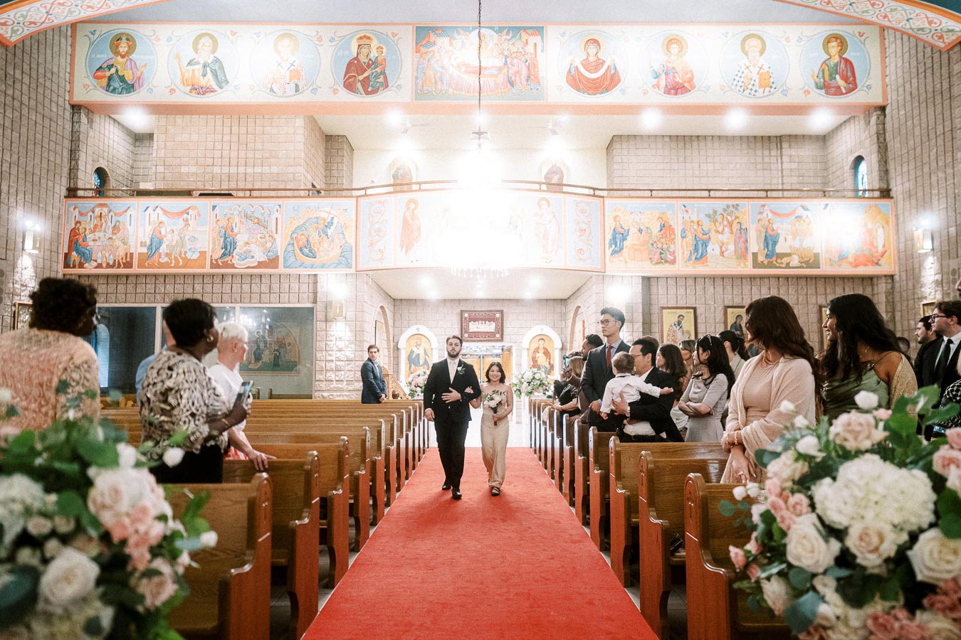 A couple walks down the aisle during a ceremony in a beautifully decorated church, featuring ornate religious artwork and a