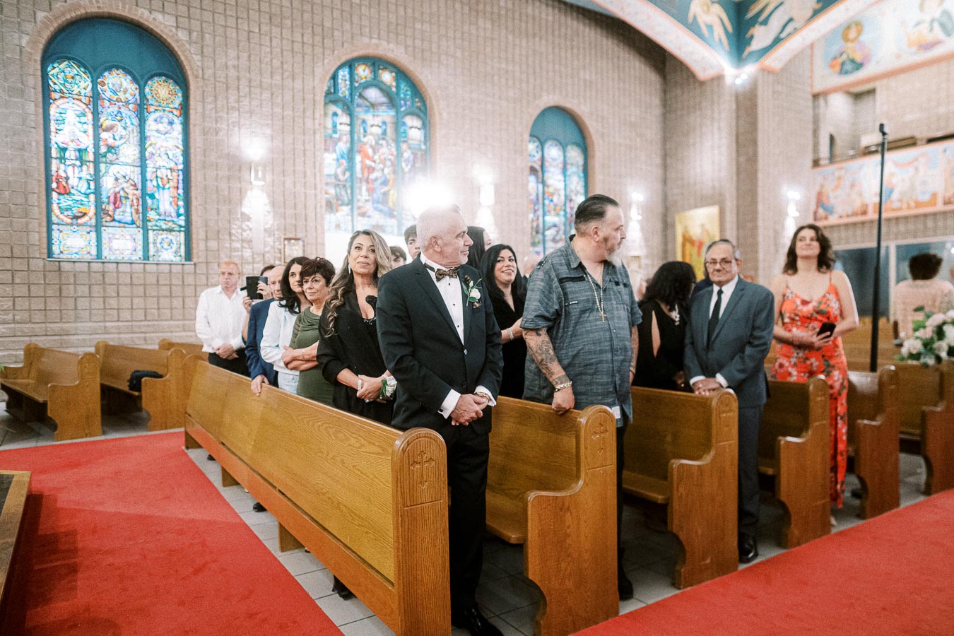 A group of people standing in a church with stained glass windows during a wedding ceremony, dressed in formal attire, with