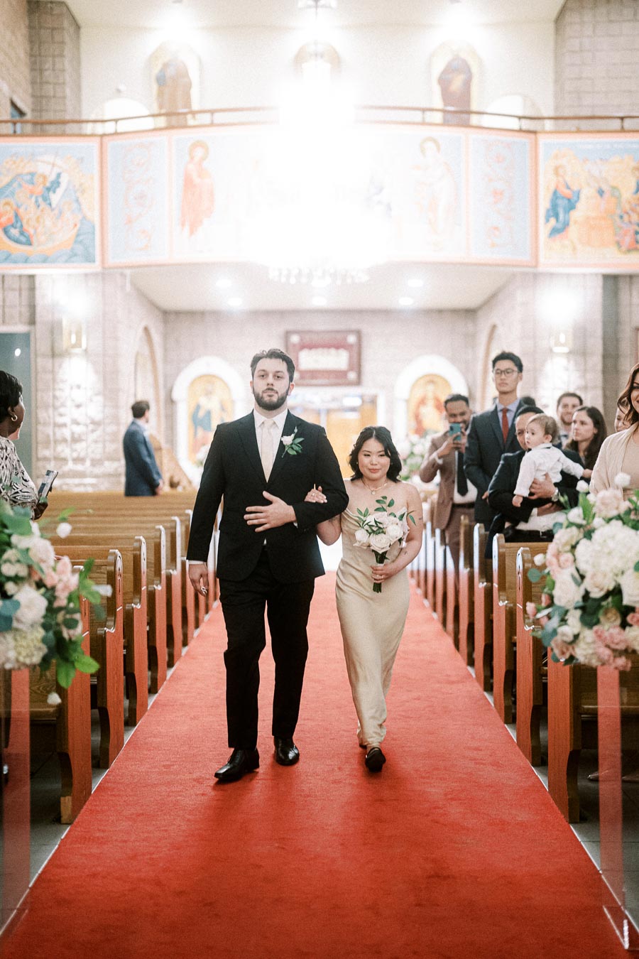 A man in a black suit and a woman in a beige dress walk down the aisle holding flowers during a wedding ceremony inside a
