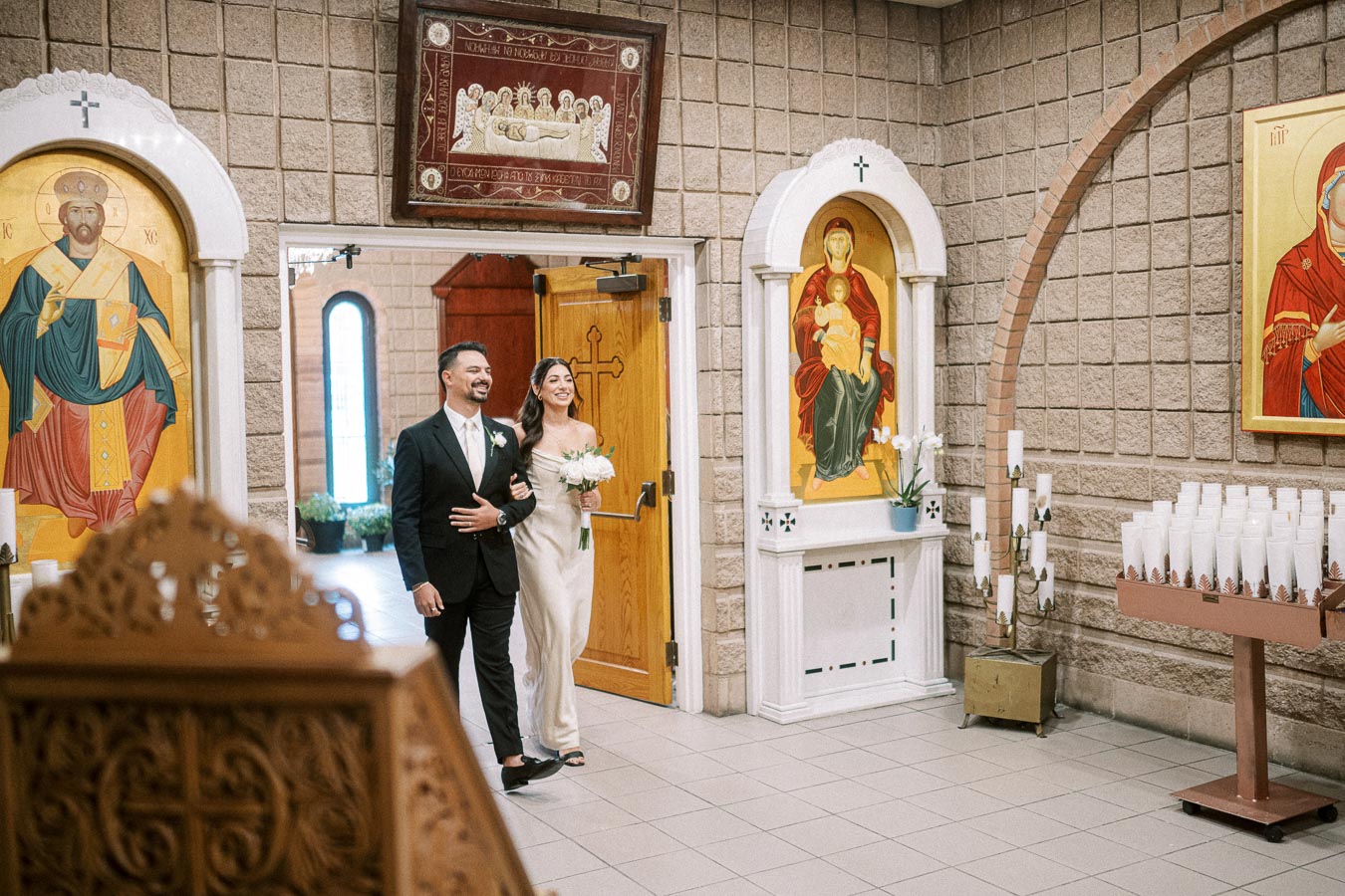A joyful couple walking arm-in-arm inside a church adorned with religious icons, holding a bouquet of white flowers, with