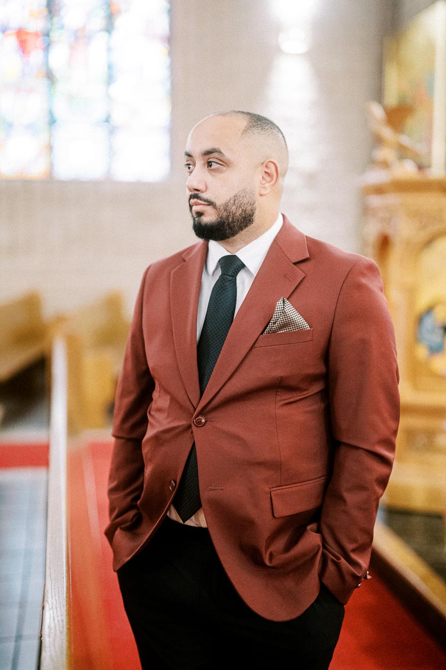 A man in a maroon suit with a black tie standing in a church with stained glass windows in the background.