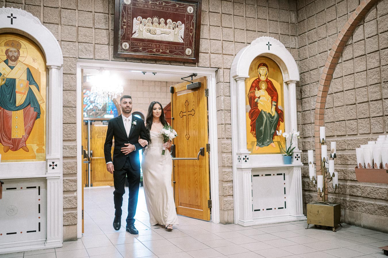 A bride and groom joyfully exit a church adorned with religious iconography and candles, signifying the commencement of