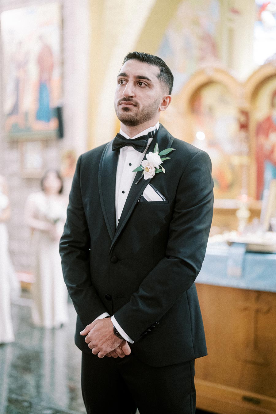 A groom in a black tuxedo with a white boutonniere stands solemnly in a church setting during a wedding ceremony.