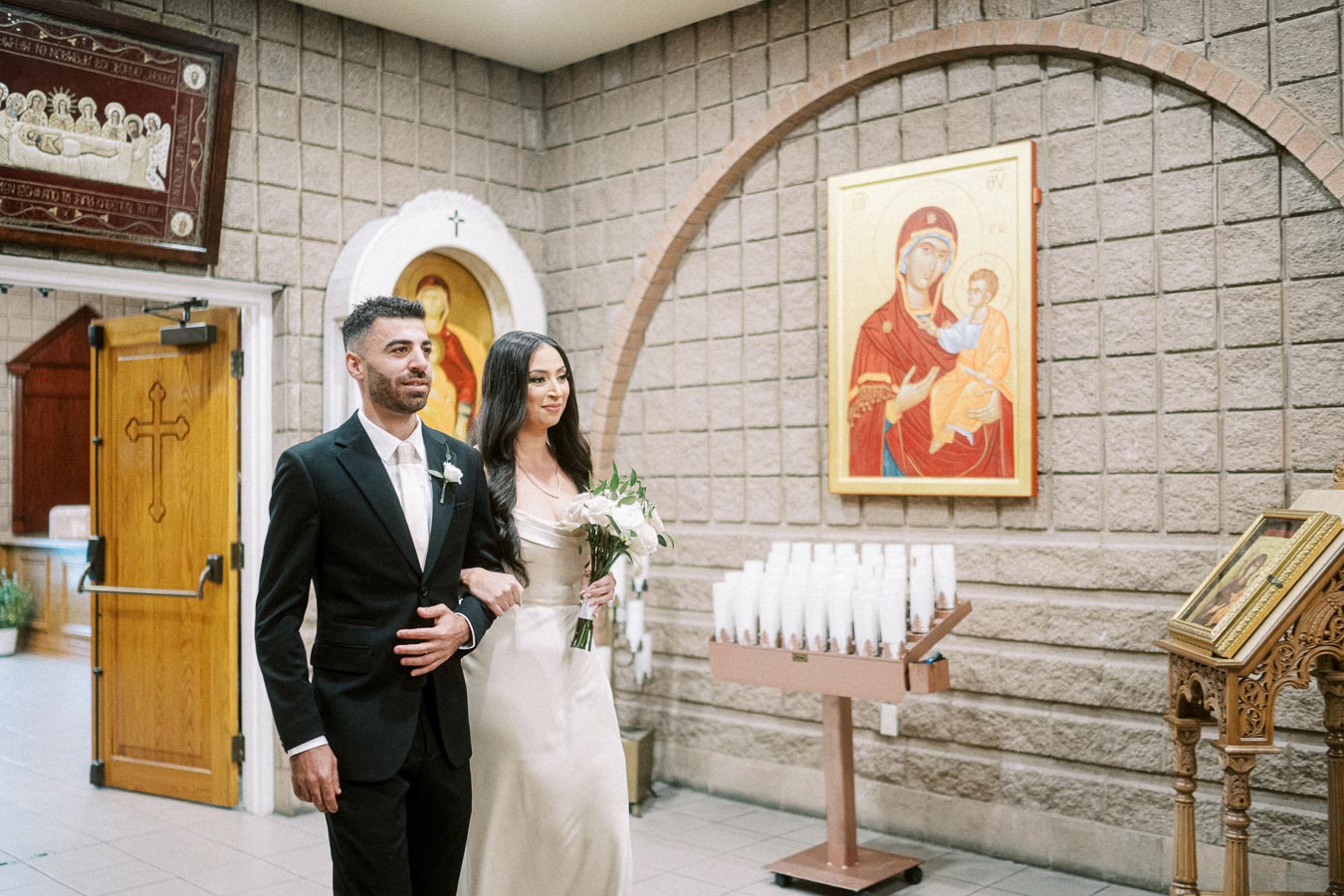 A bride and groom walking inside a church, adorned with religious icons and candles, capturing a moment from their wedding