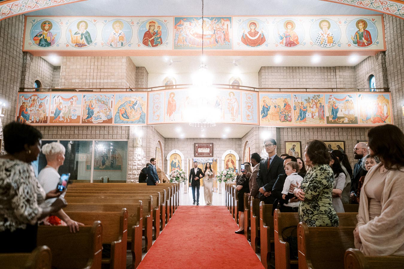 Wedding procession in a beautifully adorned church, featuring vibrant religious murals and guests lining the aisle,
