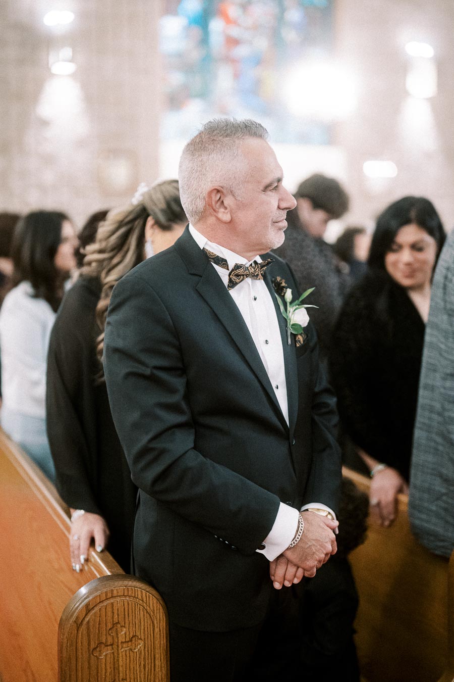 Man in formal black suit with bow tie standing in a church, surrounded by people