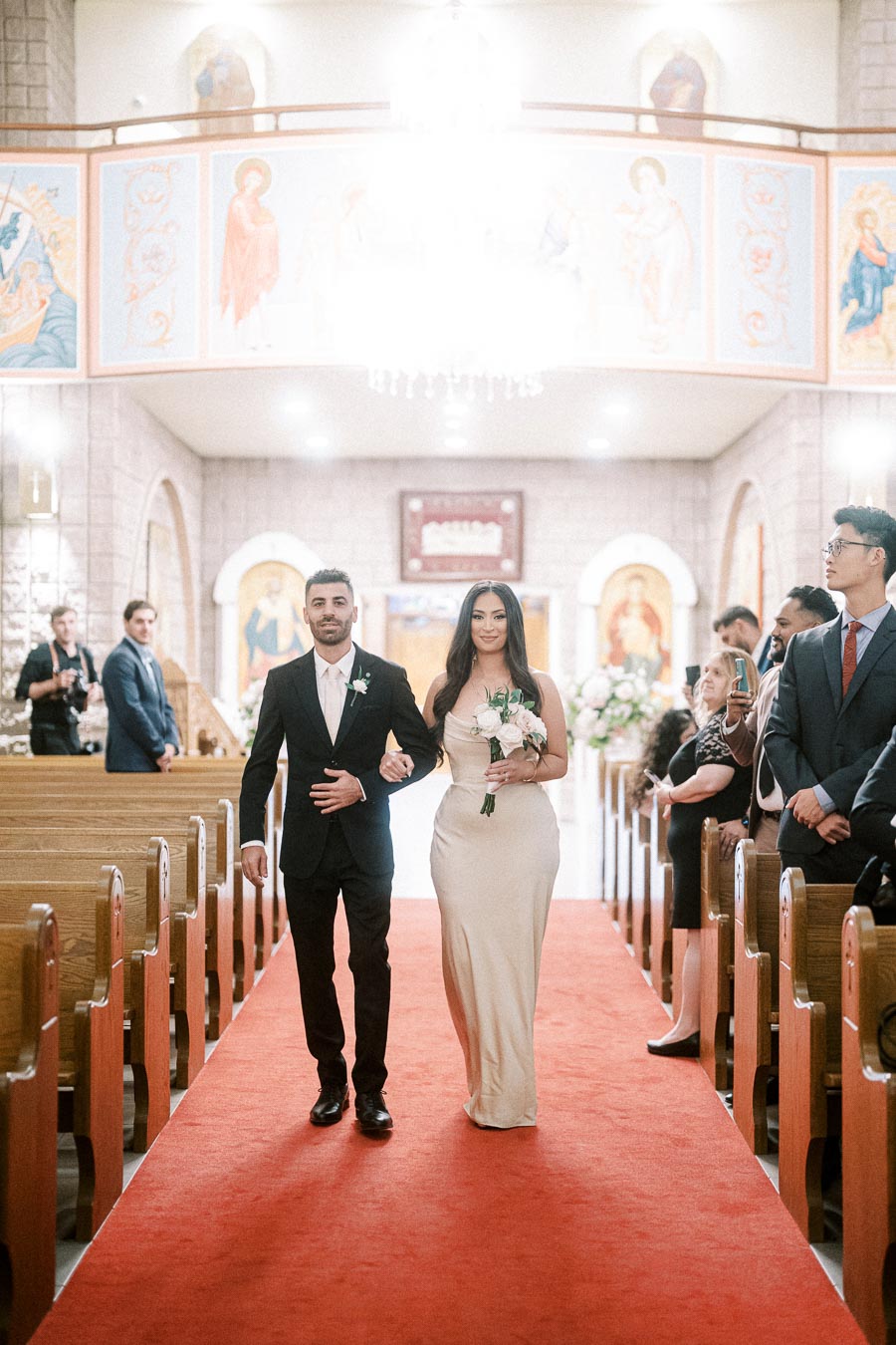 A couple walking down the aisle in a beautifully decorated church, with guests watching on both sides. The woman holds a