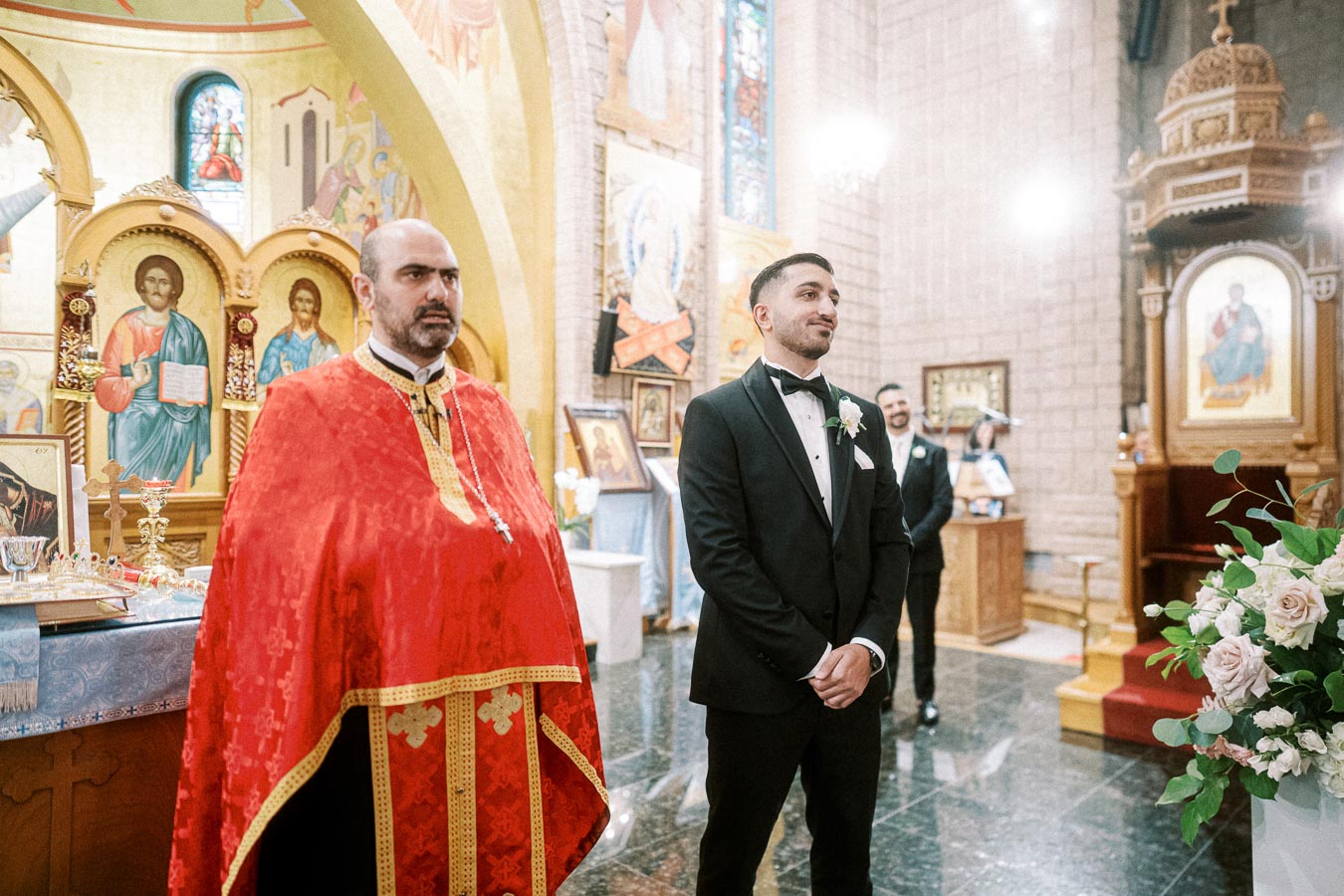 A groom in a black tuxedo stands beside a priest in a red robe inside an ornately decorated church, featuring religious