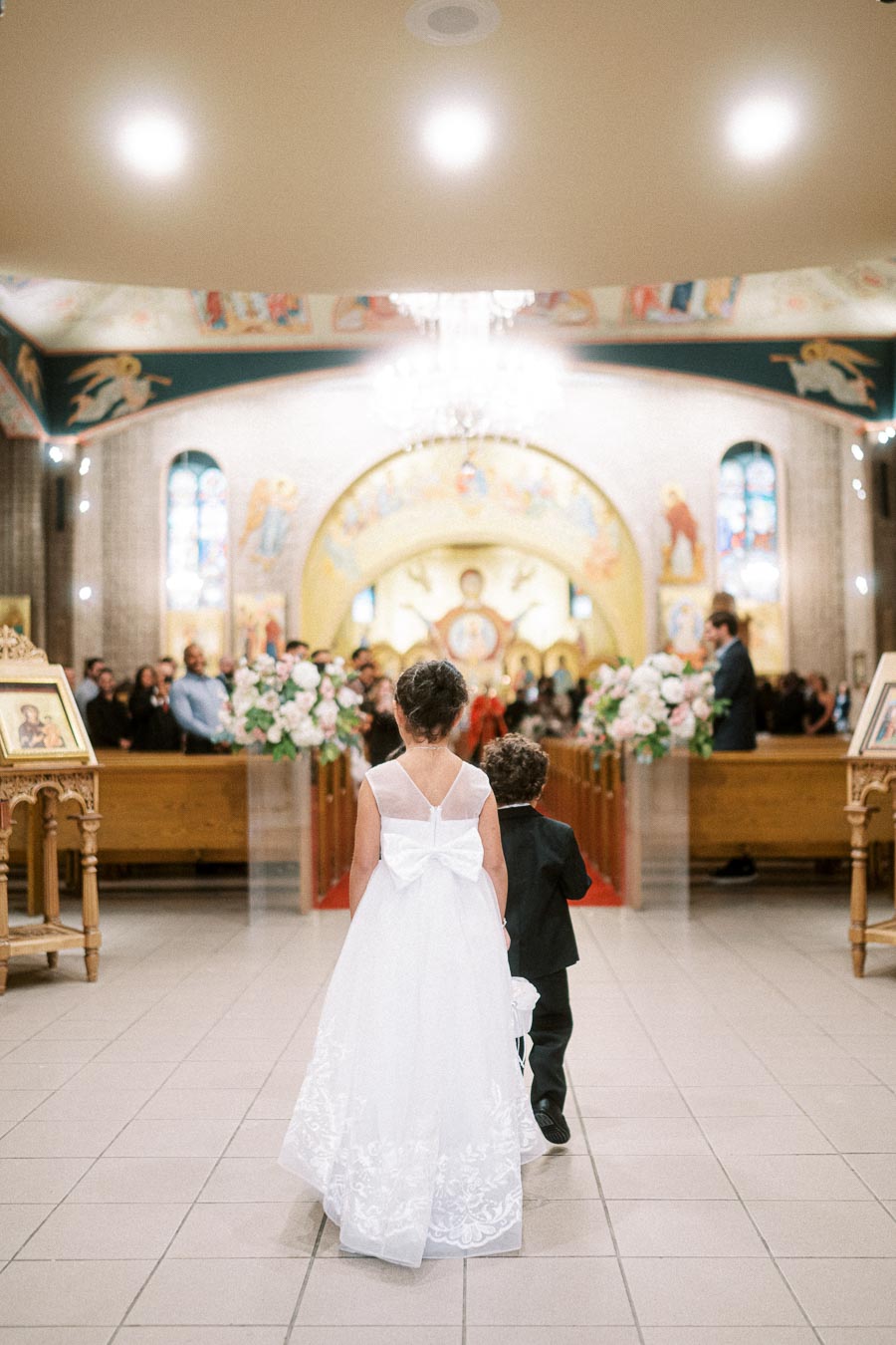 Two young children, a girl in a white dress and a boy in a suit, walk down the aisle in a beautifully decorated church