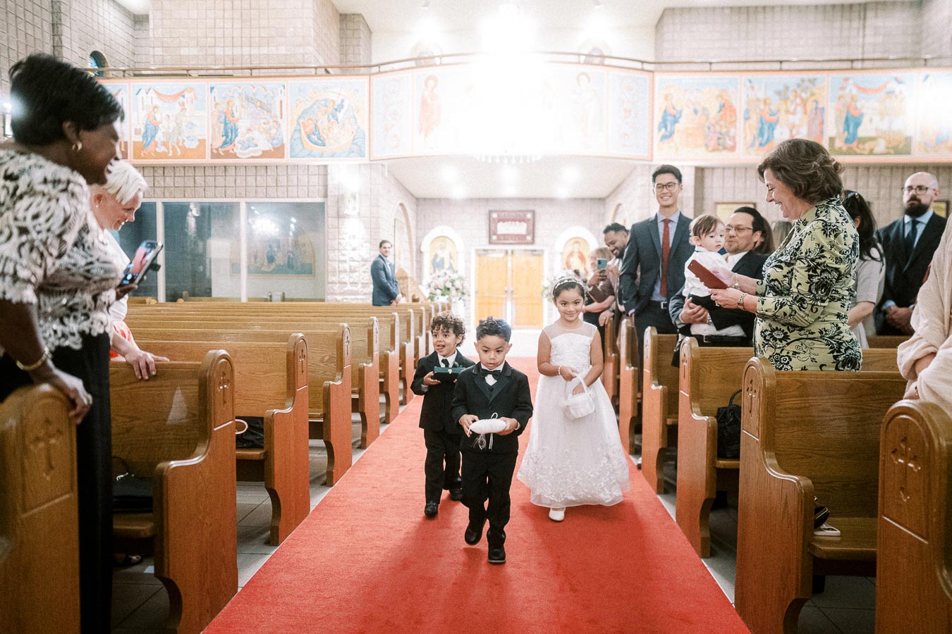 Three young children dressed in formal attire, including two boys in black suits and a girl in a white dress, walking down