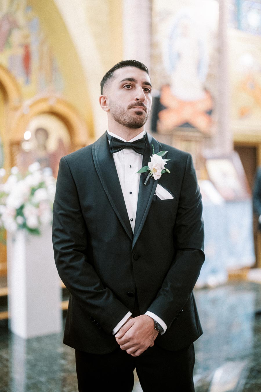 A groom in a black tuxedo and bow tie, adorned with a white boutonniere, standing in a beautifully decorated church setting,