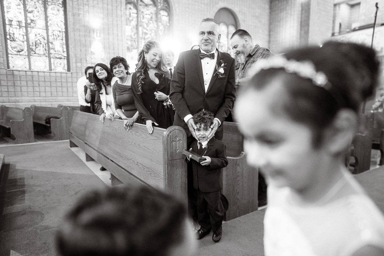 Black and white photo of a young boy in a suit standing in a church aisle in front of an adult wearing a tuxedo. The adult
