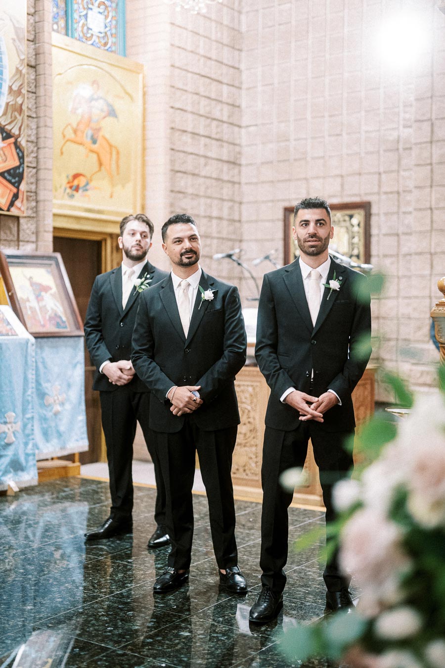 Three groomsmen in black suits and white ties standing in a decorated church, awaiting a wedding ceremony, with floral