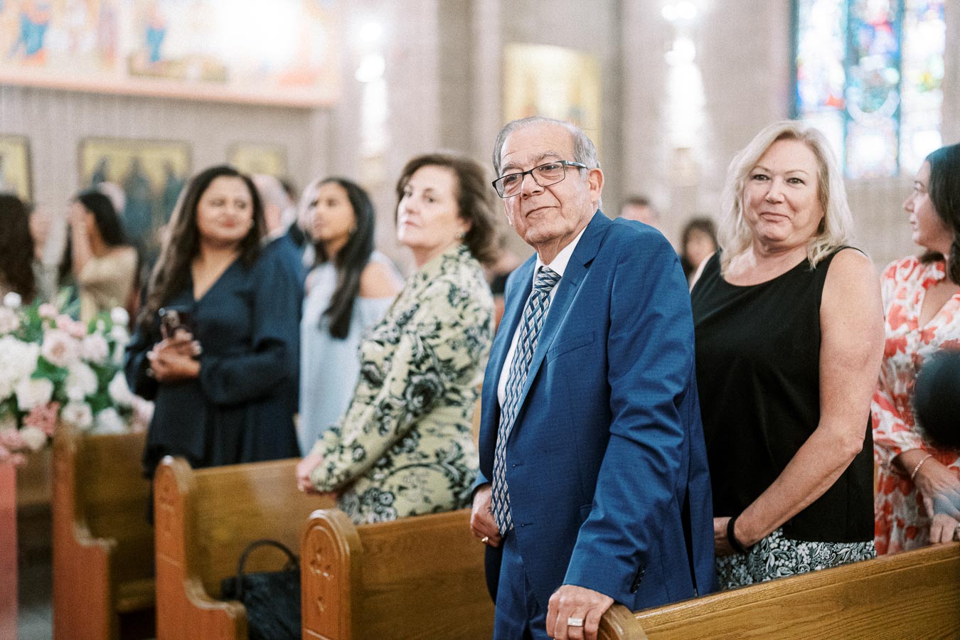 A group of people dressed formally, standing inside a church decorated with flowers and stained glass, during a ceremony or
