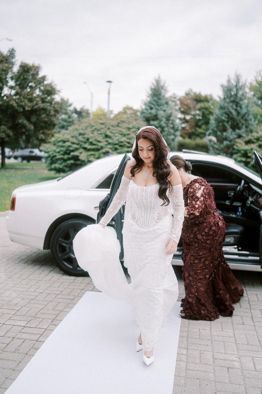 Bride in elegant white gown stepping out of a luxury car, assisted by a woman in a burgundy dress, surrounded by greenery