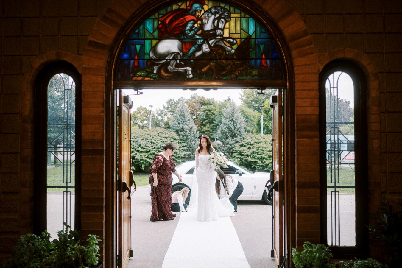 Bride in white dress entering a church adorned with stained glass, accompanied by attendants preparing for wedding ceremony,