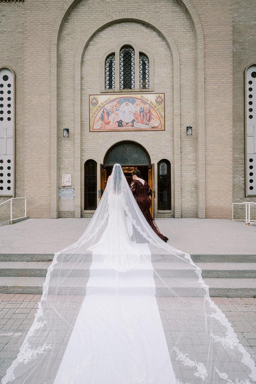 Bride in elegant white wedding gown with a long train enters a historic brick church, accompanied by another woman in a red