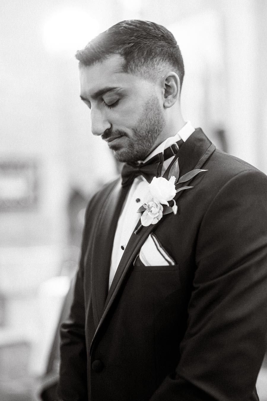 A groom in a black tuxedo with a floral boutonniere, standing thoughtfully in a blurred indoor setting.