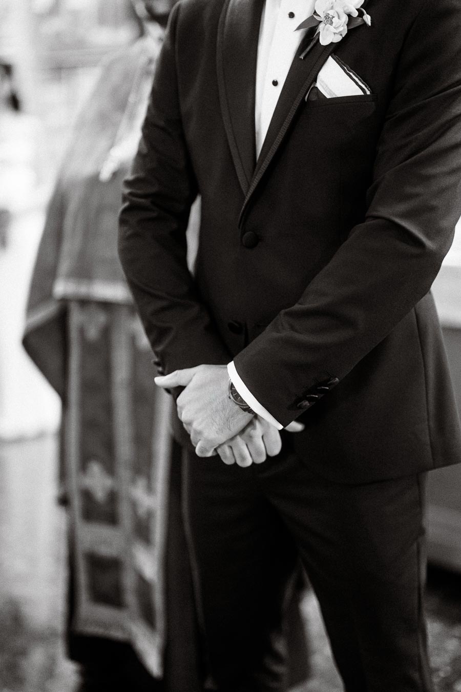 Black and white photograph of a groom in a tailored suit, standing with hands clasped at a wedding ceremony, featuring a
