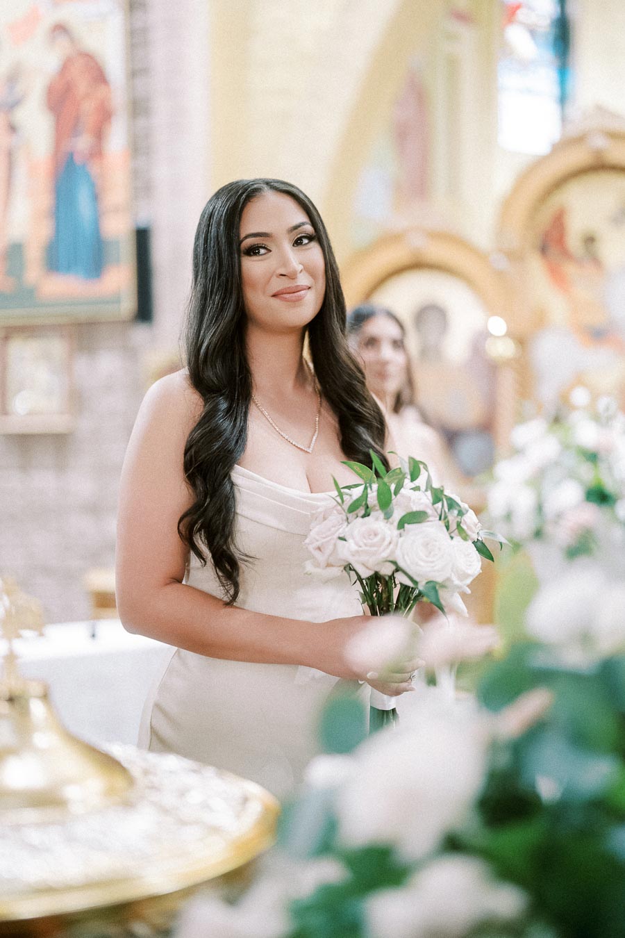 Elegant woman in a light gown holding a bouquet of roses, standing in a beautifully decorated venue with ornate artwork and